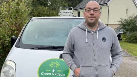 Edd Smith/BBC Ian Bowyer wearing glasses and a grey zip-up hoodie and standing in front of a white van with a Green and Clean logo on the bonnet. 