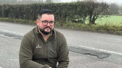 BBC A man crouching on a pavement with a repaired pothole on the road in the background