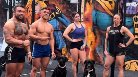 Joel McManus Two bare-chested men, one in black shorts, the other in blue shorts and two women in sports gear standing in a row with two black dogs in sitting on the ground in front of them