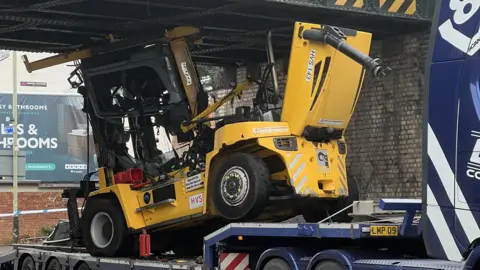 BBC The photo shows a yellow forklift, on the back of a blue lorry trailer, apparently wedged underneath a bridge. Police tape can be seen in the background, as can a billboard on the side of a building.