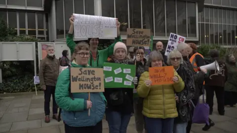 A group of people holding placards and posters, which read "save Wellington House". They are standing outside a local council building.