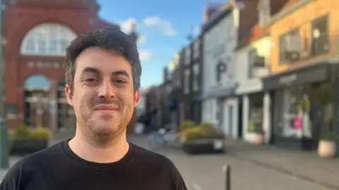 BBC News A man with brown hair and stubble, who is wearing a black T-shirt and standing in a market square with shops in the background.