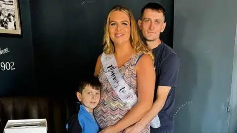 Supplied Archie York, with short brown hair, wearing a black and blue jacket, cuddles Katherine Errington's baby bump. Robbie York is to Katherine's right, holding her side.