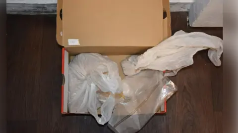Cleveland Police A cardboard shoe box open, showing white bags wrapped up inside. The box is sitting on a wooden floor in a room.