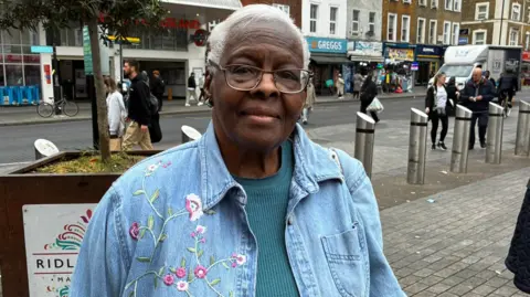 Valerie standing on a street wearing a blue denim jacket featuring a floral design. Shops like Greggs in the background.