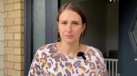 BBC A woman, Kelly Heather, wearing a colourful top and standing in the doorway of her home