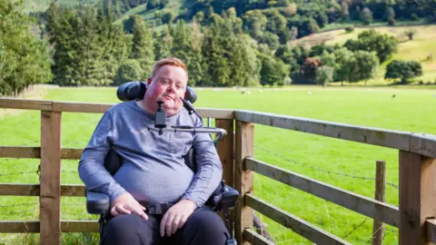 Will Clark is in his 20s or 30s with ginger hair. He is wearing a blue top and trousers and is sitting next to a fence with the trees and hills of the Lake District rising behind him. He uses a wheelchair.