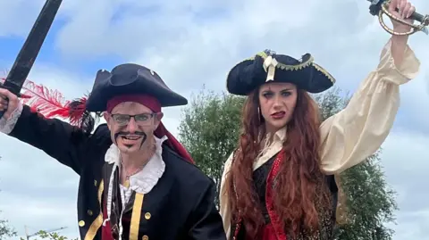 Two pirates in full costume hold two swords up aloft. 