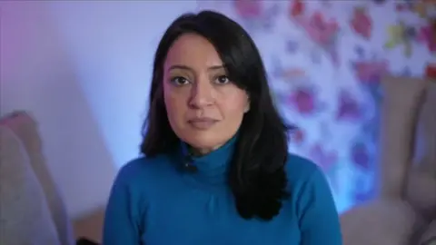 Khizra Bano, wearing a blue polo neck with long, black hair, is sitting inside with living room furniture and wallpaper visible behind her.