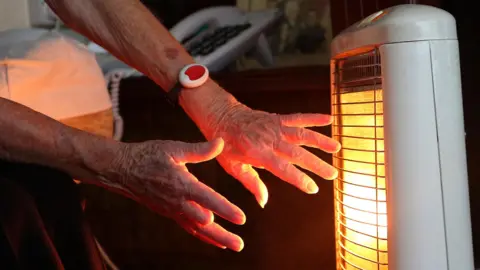 PA Media An elderly woman warms her hands at an electric fire