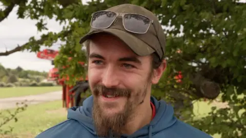 Shaun Whitmore/BBC Morgan Hermon smiles as he looks away from the camera. He is standing outside near a field. A tree can be seen behind him as well as farming equipment. He wears a green cap with sunglasses resting on it as well as a blue hoodie. He has a long beard.
