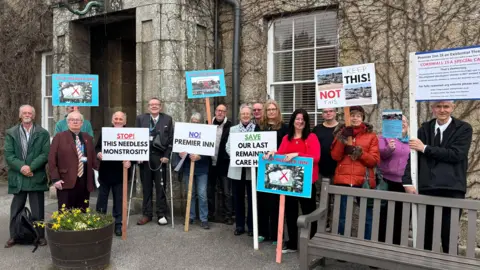 About 15 people stood outside an old looking hotel. Many of them are holding placards with phrase such as 'No Premier Inn' and 'Stop the needless monstrosity'. There is a bench to the bottom right of the photo and a flower bed to the left. 