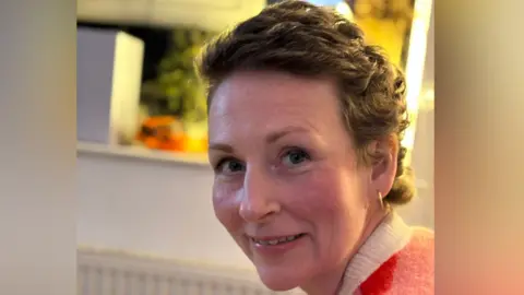 Claire is in a home and is looking over her left shoulder at the camera while smiling slightly. She has short brown wavy hair and is wearing gold hoop earrings. She has a coral coloured jumper with with a white collar and behind her is an out-of-focus kitchen.