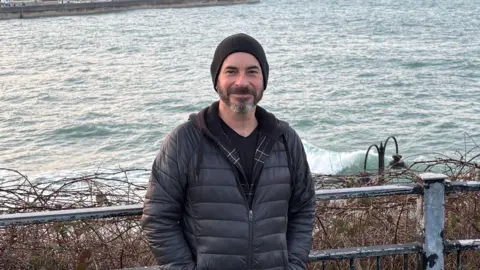 Mark wears a black beanie hat, dark t-shirt and black jacket - he stands in front of a metal fence with the sea behind him. 