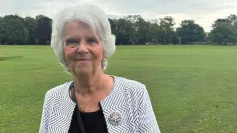 Alison Reynolds stands in the foreground of a large open grassy park, facing the camera. The person wears a black top and a light-coloured patterned jacket with a circular brooch. A wide green field extends behind them with scattered trees and a line of denser woodland in the distance. The sky is overcast with grey clouds, creating soft, diffuse lighting across the scene.