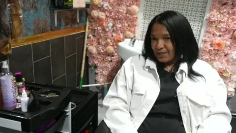 A woman sits in a salon with pink flowers behind her. Hair products can be seen on a stand to the left of her.