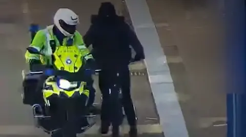 Northamptonshire Police A police officer, on a motorbike, next to a man on a bike, all dressed in black. The officer is wearing a helmet and full high-vis uniform. 