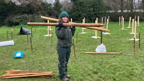 A woman stands on a patch of grass holding two large wooden stakes. Around her are lots of smaller stakes supporting newly planted trees.