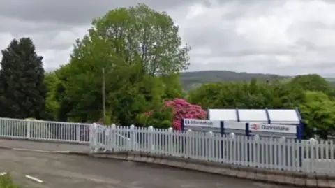 Google Street view of the Gunnislake train station. There is a white picket fence with a sign with three white sections. On the right it says information and on the left it says Gunnislake. Behind is trees with pink flowers