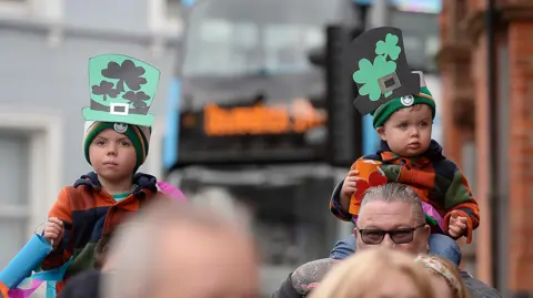 Pacemaker Young children at Downpatrick being held up on adults shoulders. Both are wearing hats with shamrocks on them. 