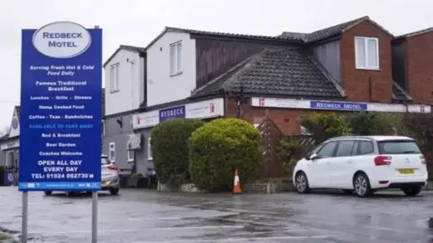 The front of the Redbeck Motel, as indicated by a blue and white sign in its car park. The sign plugs its food offering and features contact details. Two cars are parked outside the hotel, which is a relatively small two-storey building.