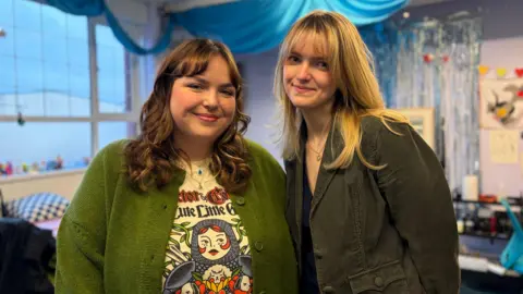 A woman with brown hair is wearing a green cardigan is stood smiling next to another woman with blonde hair and a black jacket.
