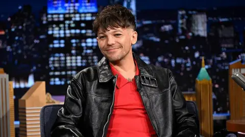 NBC via Getty Images Louis Tomlinson sat down for an interview on a US chat show. He's a white man with short dark hair and is wearing a red T-shirt with a black leather jacket. He is smiling with his mouth closed and behind him there is a backdrop that looks like a US cityscape.
