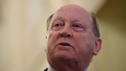 A balding dark haired man with blue eyes looks to the left of the camera. The background is blurred.
