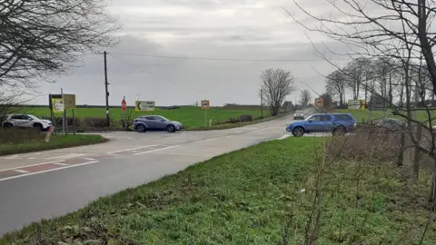 A few cars getting ready to drive through a cross roads. There are fields all around the roads. It is an overcast and grey day.