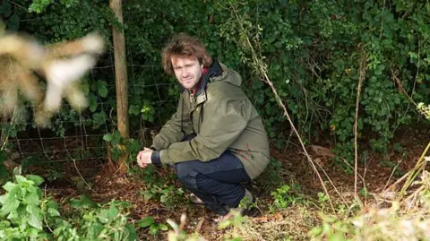 BBC/Wall to Wall Media/Stephen Hart Una persona con una chaqueta verde oliva para actividades al aire libre se agacha junto a una valla de alambre al borde de un denso bosque. El suelo está cubierto de hojas, hierba y vegetación baja, mientras que un espeso follaje verde llena el fondo. Plantas ligeramente desenfocadas enmarcan el primer plano, resaltando el entorno rural aislado.