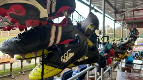 Close up   changeable  of galore  pairs of rugby boots hanging connected  a line. There are a assortment  of colours though  astir   of them are achromatic  boots with assorted  sports marque  logos connected  them. In the inheritance  there's a rugby transportation   and immoderate   scaffolding poles are besides  visible.