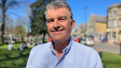 Andrew Lund-Yates. He has short grey hair, no facial hair and is wearing a blue and white pinstripe shirt. He is looking at the camera, squinting in the sun and smiling
