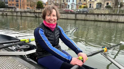 Katherine Webster is sitting in a boat on the river, smiling at the camera. She has short brown hair and is wearing a pink scarf and a black and blue jacket and trousers. There are houses, flats and parked cars on the road on the other side of the river behind her