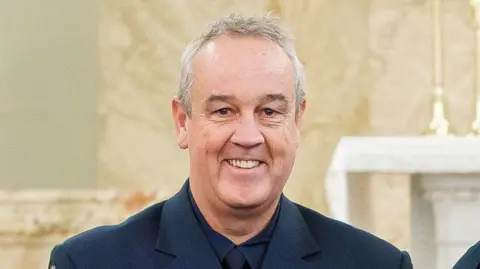 A middle-aged man with grey hair, smiling at the camera and wearing a dark blue suit