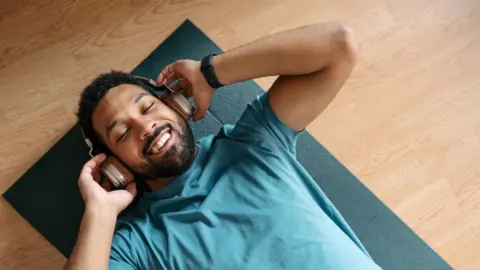 Getty Images Man wearing blue t-shirt, lying on a dark green yoga mat on a wooden floor, with over-ear headphones on. He's smiling and has his eyes closed.