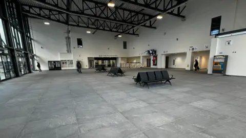 Sunderland Station's southern entrance. The area is sparsely filled with just a few seats, a ticket machine and vending machines in the distance. The flooring is grey, while the walls are painted white.