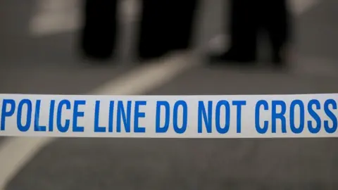 A stock image of white police tape across a road with blue writing that reads 'police line do not cross'. The background is blurred.