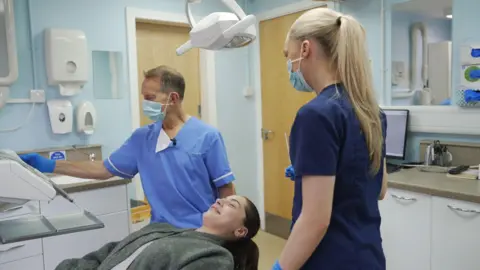 BBC Glasgow dentist David McColl with a patient undergoing a check-up - she is lying down and smiling. He is in his medical scrubs and checking a machine. A dental nurse is standing next to the patient, with her back to the camera.   