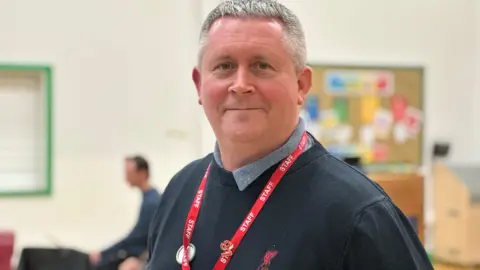 Juan Garrett, who short grey hair, standing in a school hall during a dance rehearsal. He is wearing a dark blue jumper and has a red lanyard with staff written on it around his neck.
