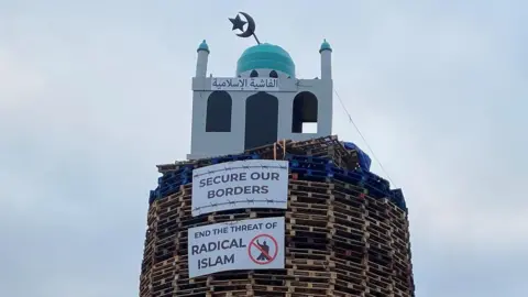 BBC An image of the top of the bonfire, consisting of wood pallets, with a structure resembling a mosque on top. Two placards are below. One reads "secure our borders" while another reads "end the threat of radical Islam".