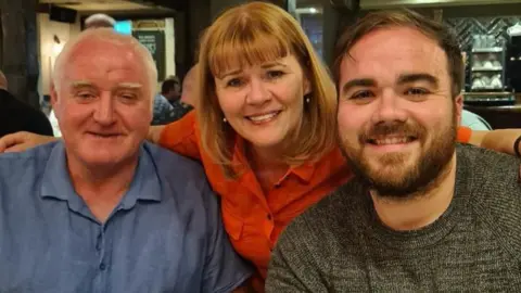 PA Elena Walsh, a 64-year-old woman with short ginger hair and wearing a red shirt, smiles with her arms around her husband, a main with grey hair and a blue shirt, and her son, a younger man with short brown hair, a beard and a grey jumper. 