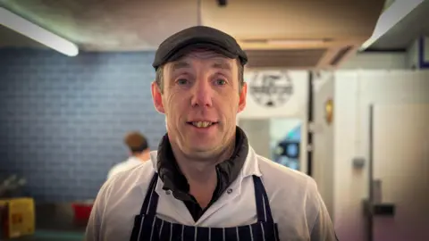 Aled is standing in a butchers shop looking at the camera. he is wearing an apron and while smock with a black flat cap. 