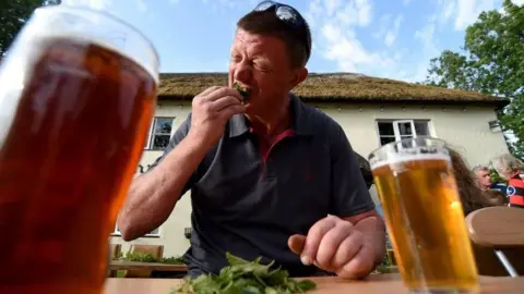 A man sitting at a table outside the Bottle Inn. He is pushing a handful of stinging nettle leaves in his mouth and pulling a pained expression. On the table in the foreground are two full pints of beer or cider.