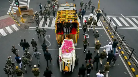 AFP via Getty Images The funeral procession shows several people walking behind a coffin covered in colorful flowers on a New Delhi street