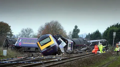 The wreckage of a blue and yellow train by the side of a railway, with workmen in high-visibility clothing milling around next to it.