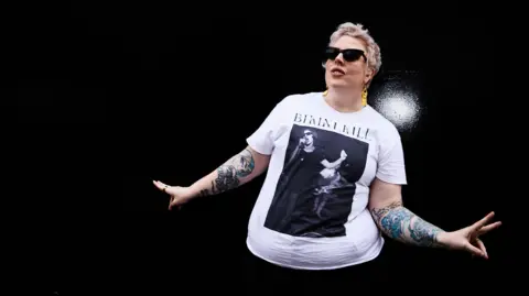 Eva Pentel Marea Stamper, alias La Sainte Madone, porte une paire de lunettes de soleil foncées et un t-shirt blanc, avec les tatouages ​​sur ses bras exposés