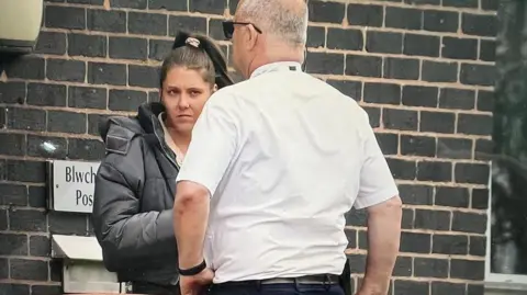 Shannon Ives is pictured on the left in a black jacket and black jeans with her hair in ponytail, talking outside Mold Crown Court with a man dressed in white shirt and black trousers.