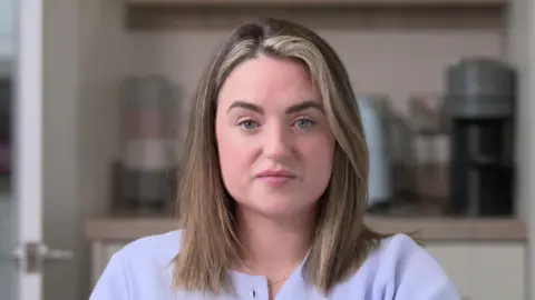 Alyssa McCrea is sitting in a kitchen. She has shoulder length fair hair. She's wearing a lilac cardigan and a silver necklace.  