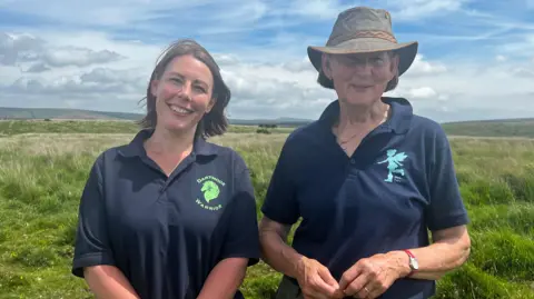 Jo Lovemore and Sheila Robson are standing next to each other in open moorland. Grassland is visible behind them with some hills in the distance. It is a sunny day but the sky has some clouds. Jo and Sheila are both wearing navy polo tops.