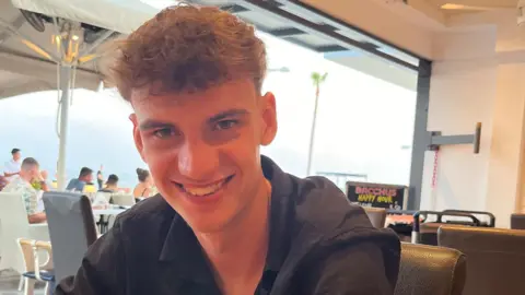 Zain Shaw, a young man with curly brown hair and a black shirt on, smiling at the camera, while sat in a restaurant.